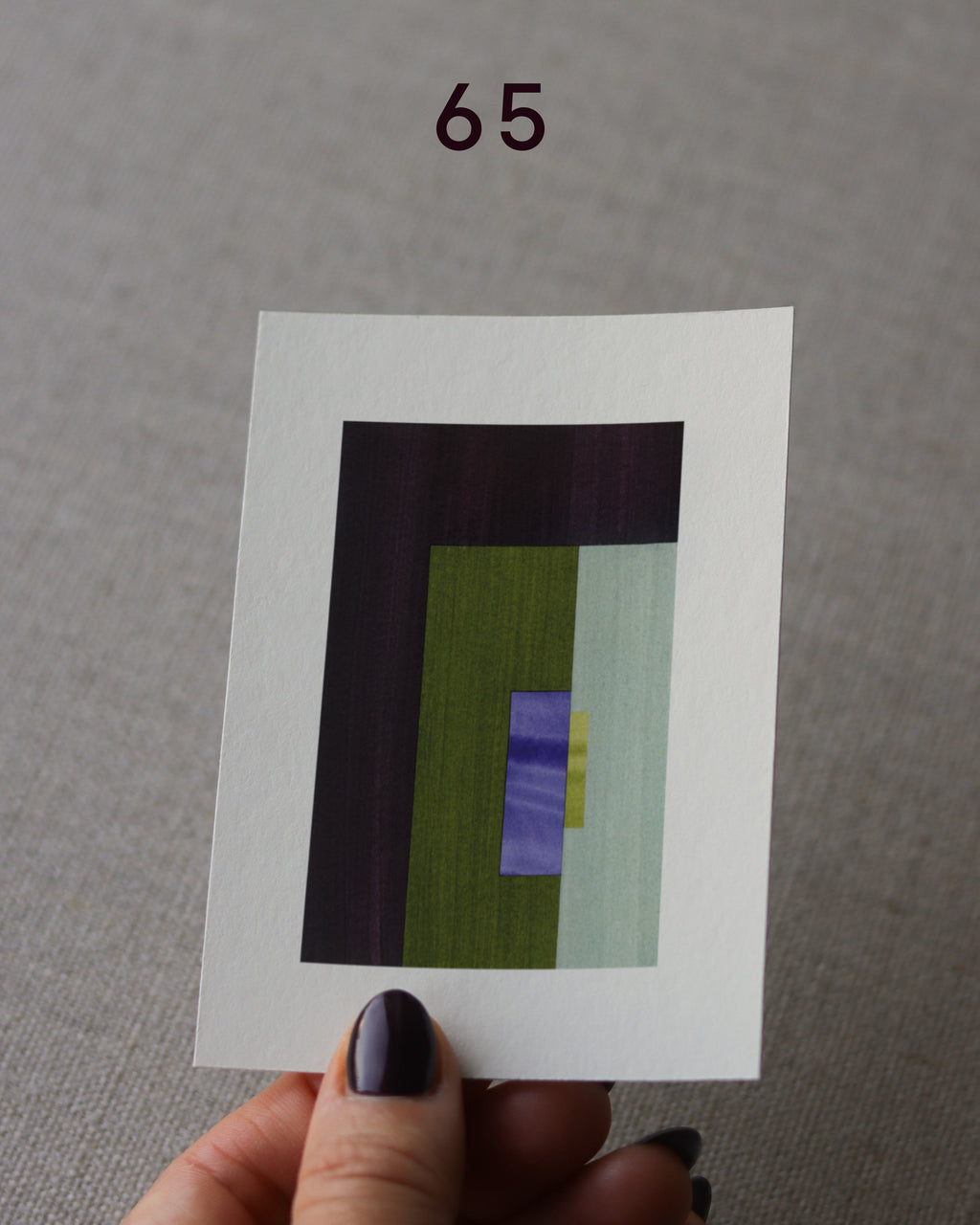 Hand holding sample of an olive green, dark brown and blue, minimalist abstract art print with a geometric design over a grey linen background