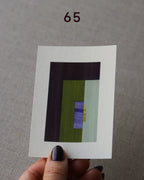Hand holding sample of an olive green, dark brown and blue, minimalist abstract art print with a geometric design over a grey linen background