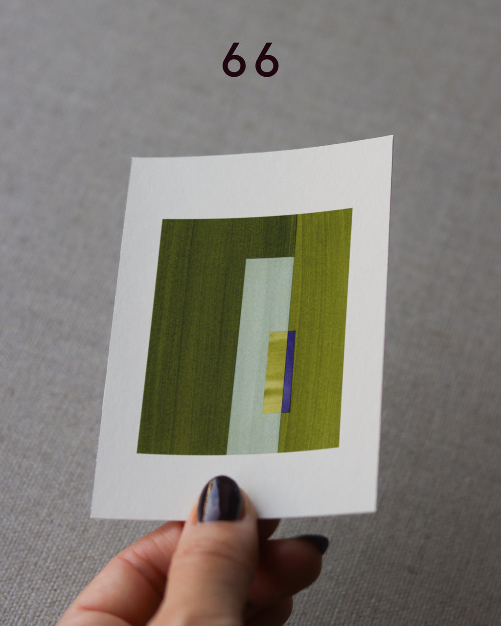 Hand holding sample of a green and purple minimalist abstract art print with a geometric design over a grey linen background