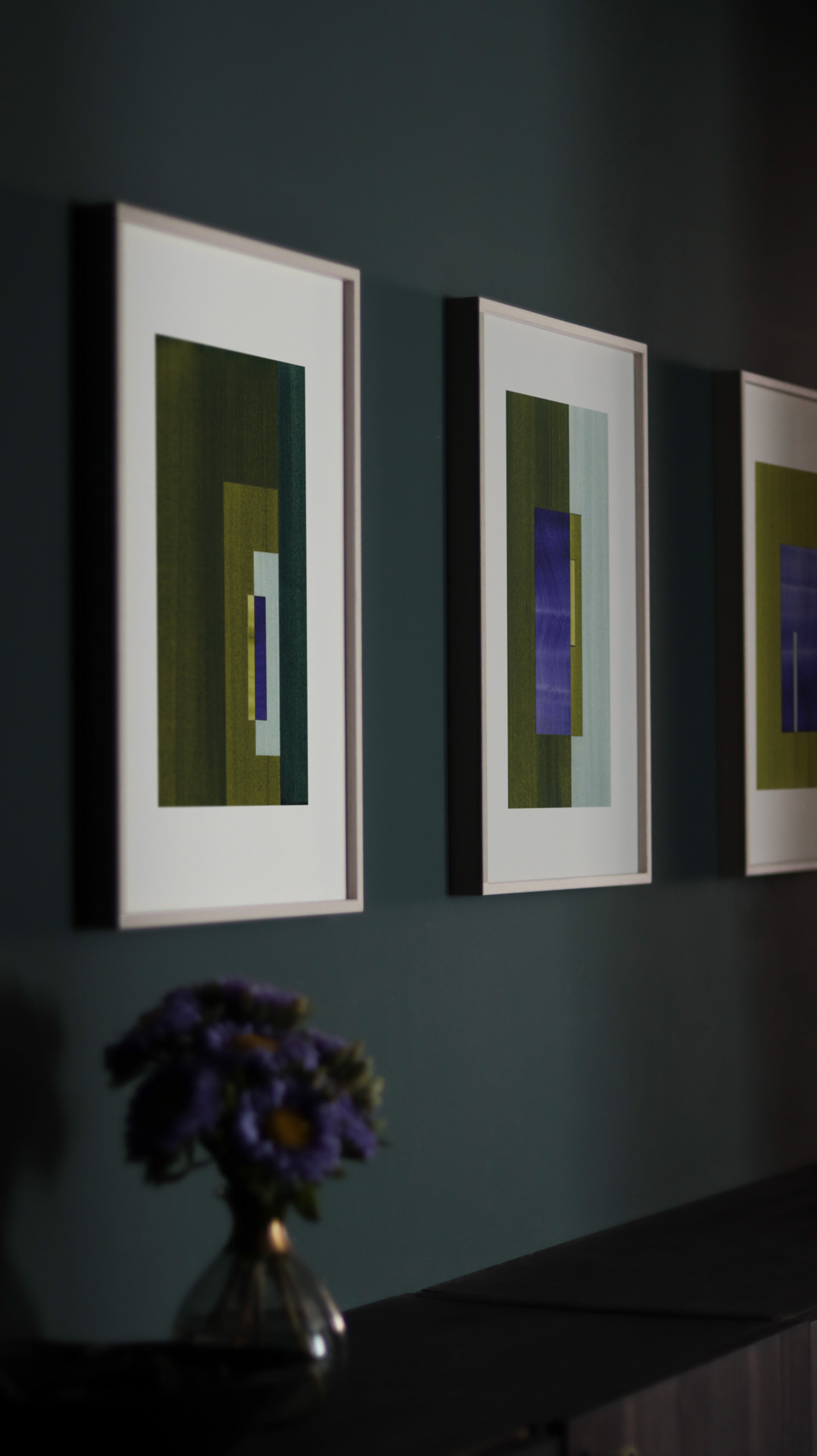 Blue and green geometric abstract art prints framed in white oak frames, hung in a moody dark green hallway. Dark interior inspiration.