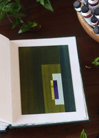 Open sketchbook with a green geometric abstract art piece on a dark wooden table with tubes of watercolour paint and plants.