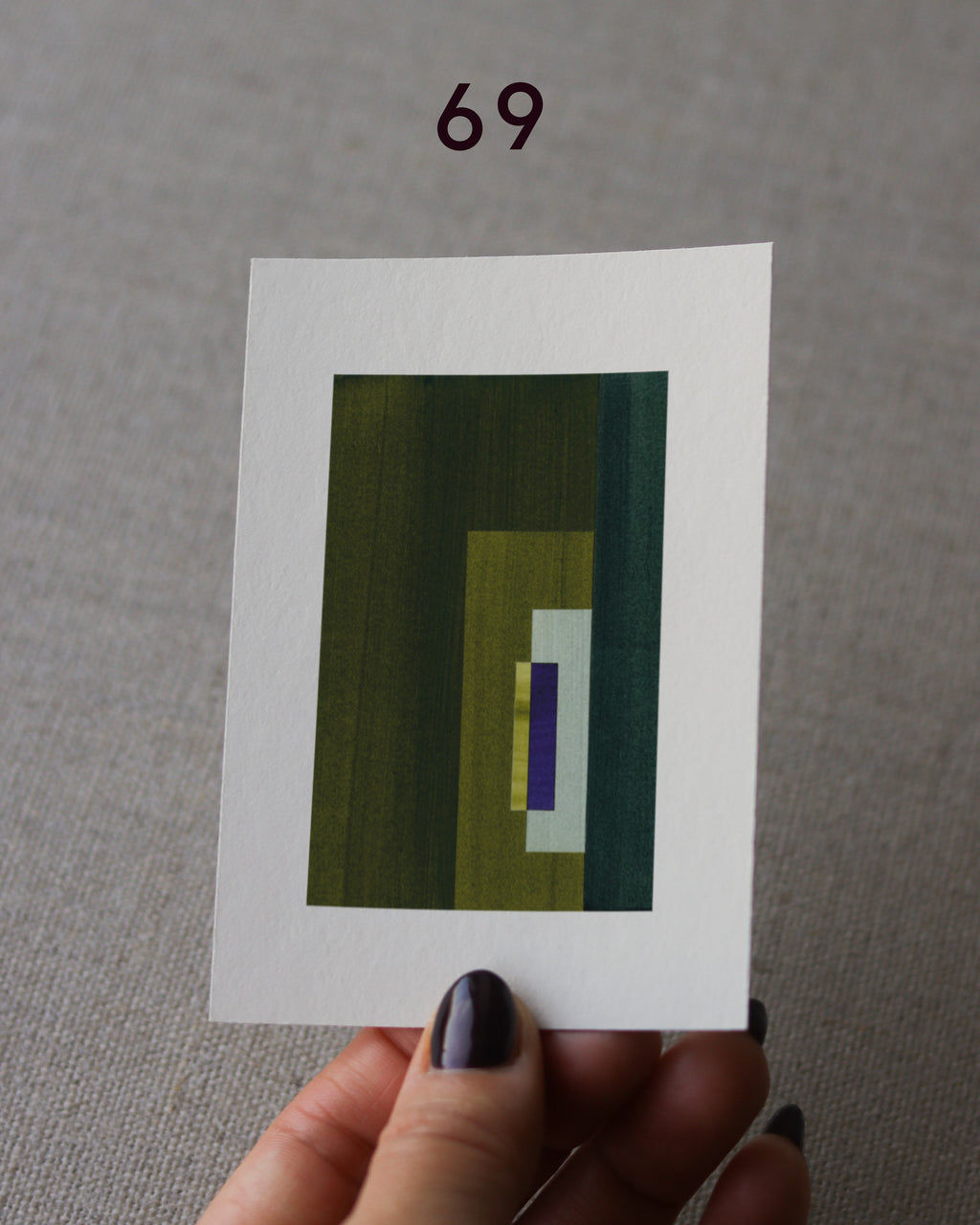Hand holding sample of an olive green and royal blue minimalist abstract art print with a geometric design over a grey linen background