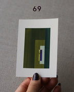 Hand holding sample of an olive green and royal blue minimalist abstract art print with a geometric design over a grey linen background