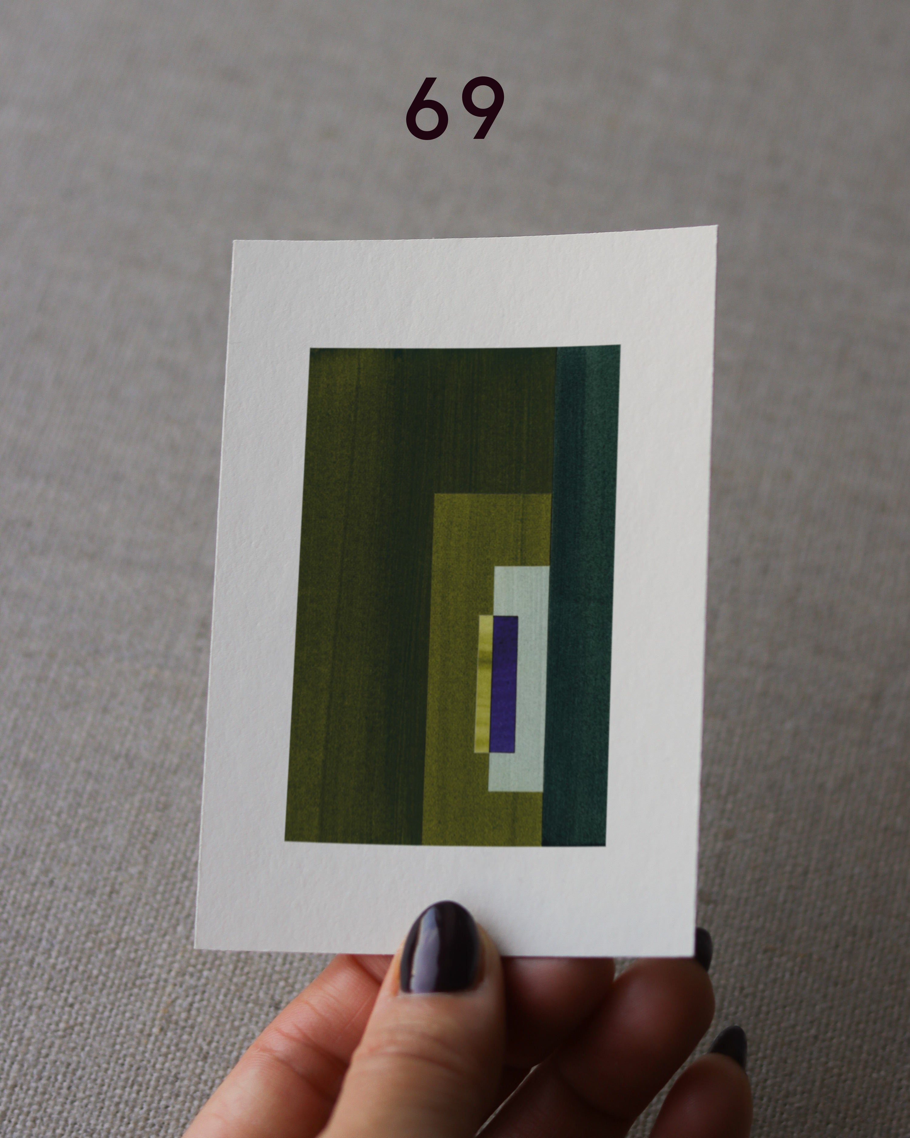 Hand holding sample of an olive green and royal blue minimalist abstract art print with a geometric design over a grey linen background