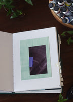 Open sketchbook with a turquoise and purple geometric abstract art piece on a dark wooden table with tubes of watercolour paint and plants.